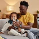 An African American man helps a child use an inhaler. They are sitting on a couch in a casual home setting. 