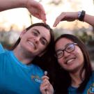 Two students wearing blue shirts smile and form a heart with their arms.