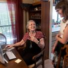 An older woman sitting in front of a home computer and a clinical neuropsychologist, who is standing, talk