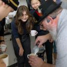 Students gather and watch with excitement as a white-bearded man in a light gray shirt, glasses, black open-ear Bluetooth headphones and a feathered black fedora pours foam into a small, white coffee mug.