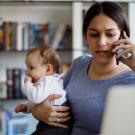 Woman holding small child while talking on phone. They sit at desk in front of a laptop screen.
